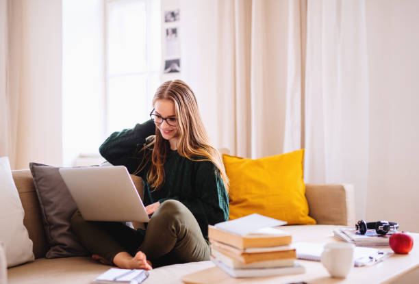 A young happy college female student sitting on sofa at home, using laptop when studying.